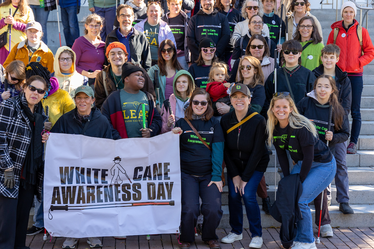 A large, cheerful group gathers on a staircase for a group photo during a White Cane Awareness Day event. Many participants wear matching black event T-shirts, and several hold white canes, showing solidarity and support. At the front, a banner reading 