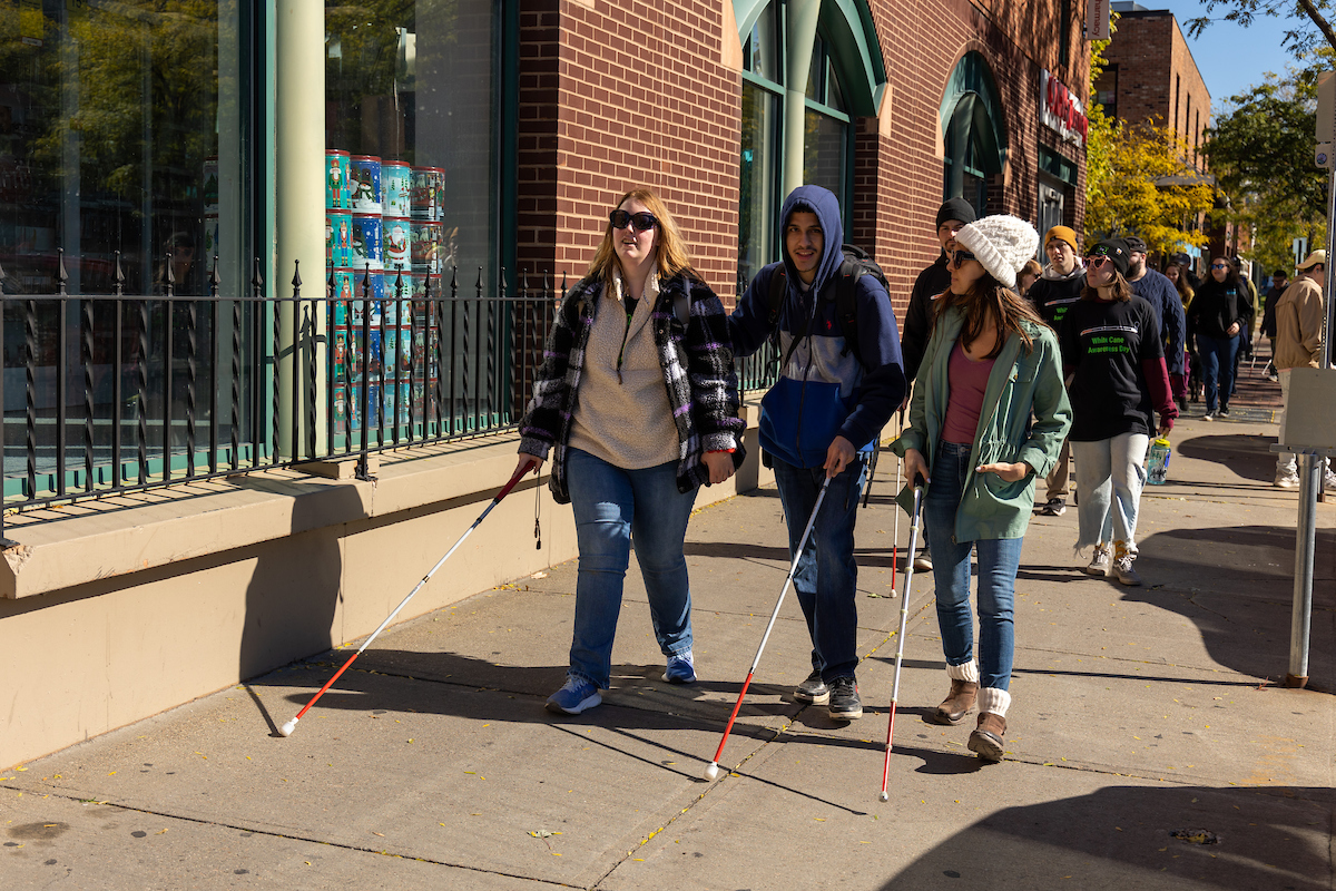 Three young adults walk side by side using white canes during a White Cane Awareness Day event. They are smiling and talking while walking past a shop window on a sunny day. A crowd of participants follows behind them along the sidewalk.